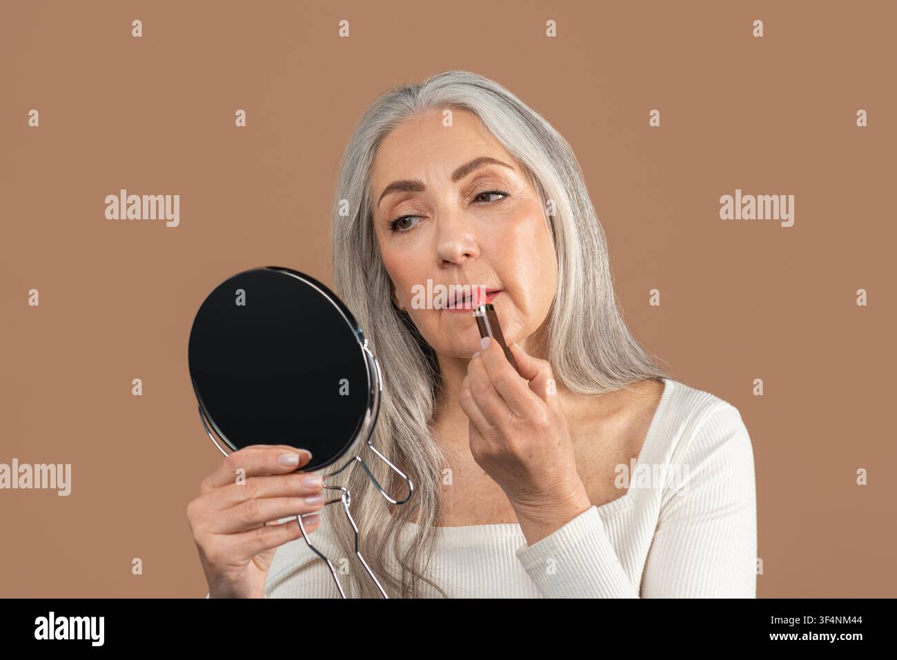 Seniorin, die Lippenstift aufträgt, während sie zu Hause in den Spiegel schaut, Schönheitsroutine Stockfoto