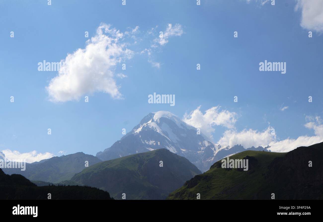 Panoramatische Ansicht des schneebedeckten Berges Kazbegi unter einem weiten blauen Himmel mit leichten Wolken und grünen Bergsilhouetten im Vordergrund, Georgien Stockfoto