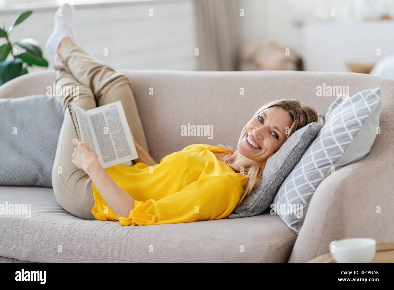 Lächelnde junge Frau liest ein Buch und entspannt sich auf der Couch in einem modernen Wohnzimmer, Wohnkonzept Stockfoto