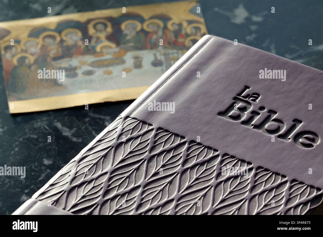 Eine französische Ausgabe der Bibel mit einem geprägten Blattmuster auf dem Einband liegt neben einem verschwommenen Bild des Letzten Abendmahls. Stockfoto