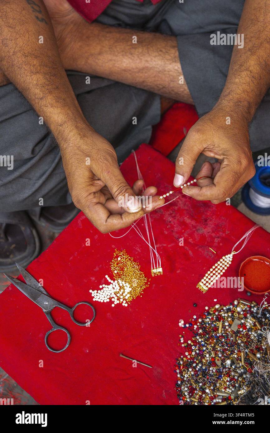 Ein Handwerker in Kalkutta, West Bengal, Indien, fädelt sorgfältig Perlen auf, um ein Schmuckstück zu erstellen, wobei er Scheren und Faden verwendet, um präzise Details zu erreichen Stockfoto