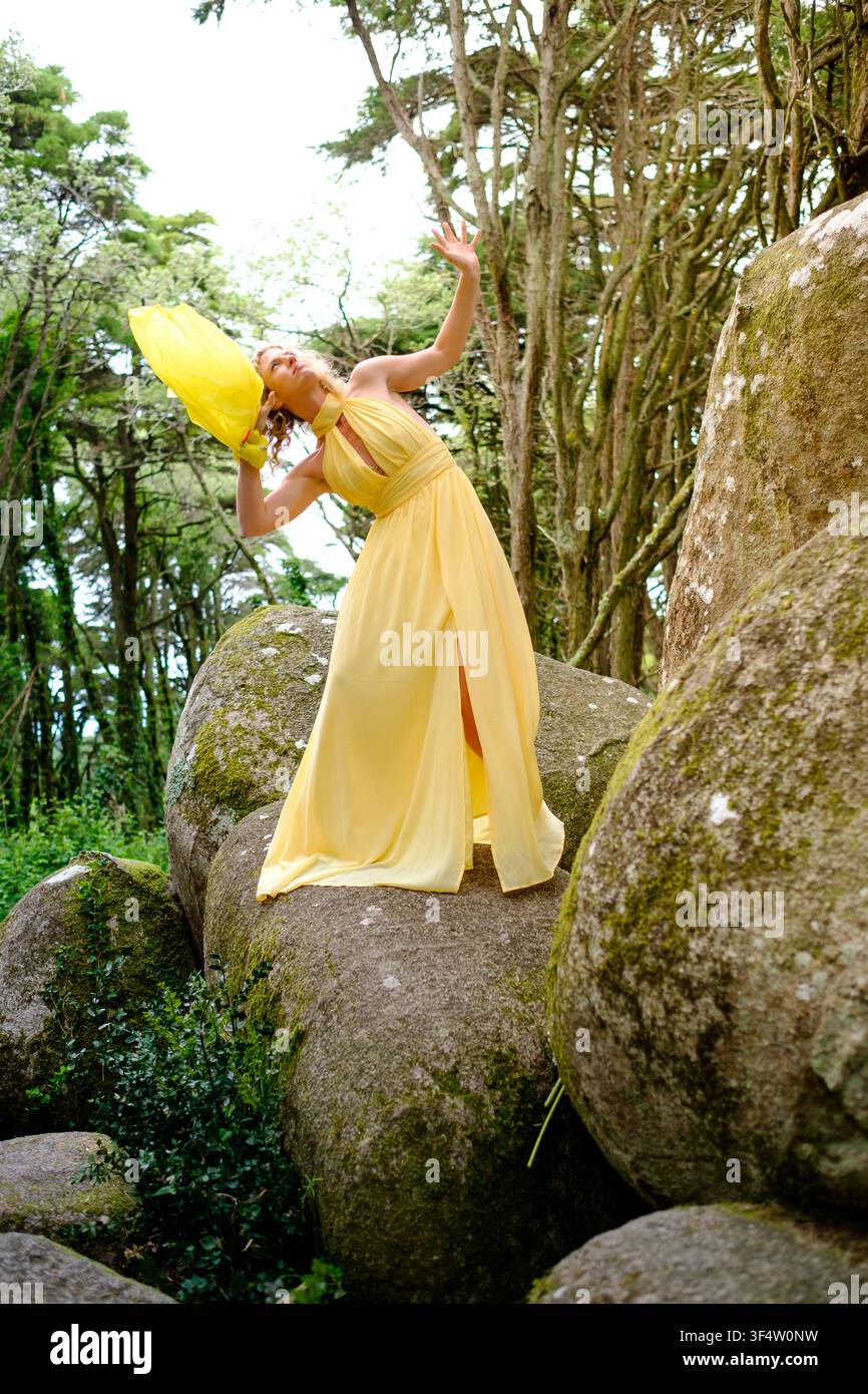 Frau in einem fließenden gelben Kleid, die auf großen moosbewachsenen Felsen in einem grünen Wald steht Stockfoto