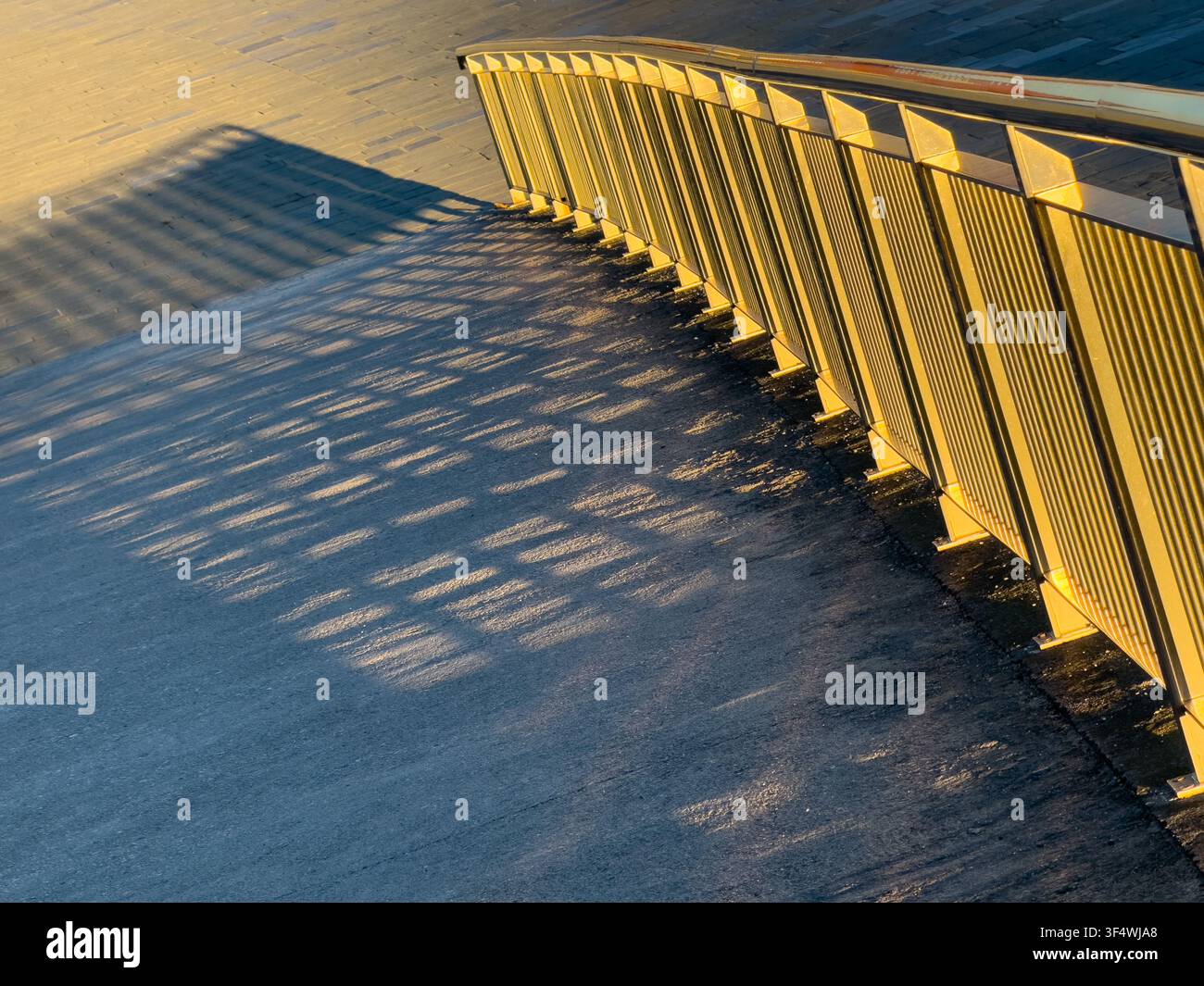 Goldenes Sonnenlicht wirft sich wiederholende Geländer-Schatten auf den Bürgersteig in einer warmen urbanen geometrischen Komposition. Stockfoto