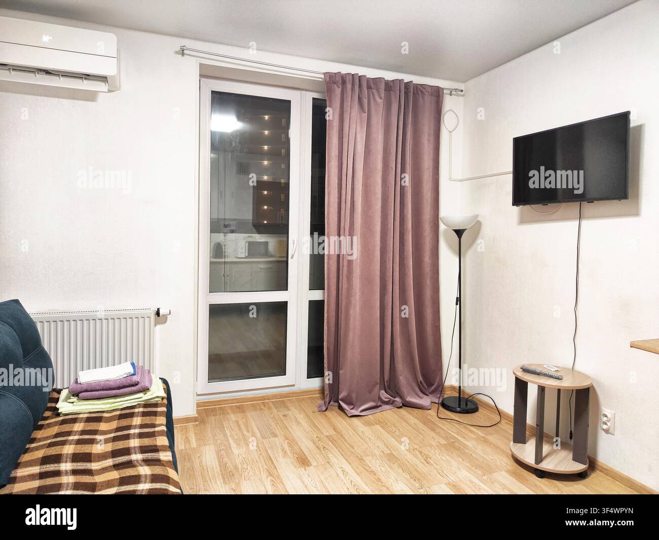 In einem Wohnzimmer steht eine Couch neben einer Lampe und ein Fernseher hängt an der Wand, während Vorhänge das Fenster bedecken. Stockfoto