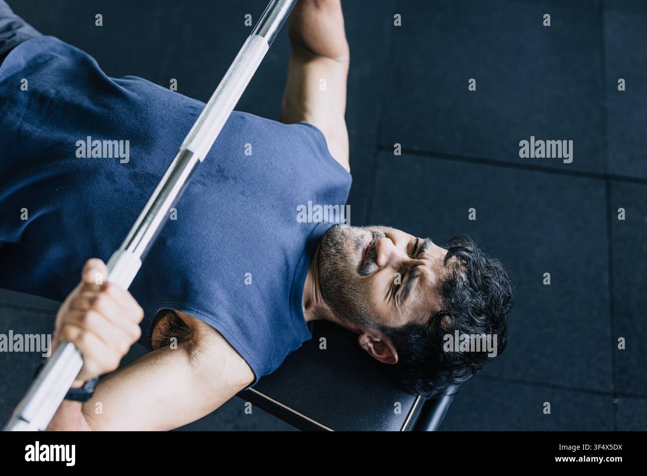 Brustmuskelaufbau beim Bankdrücken, Nahaufnahme eines sportlichen Mannes, der in liegender Position eine schwere Hantelstange hebt, mit sehr intensivem Gesichtsausdruck im Fitnessstudio. Stockfoto