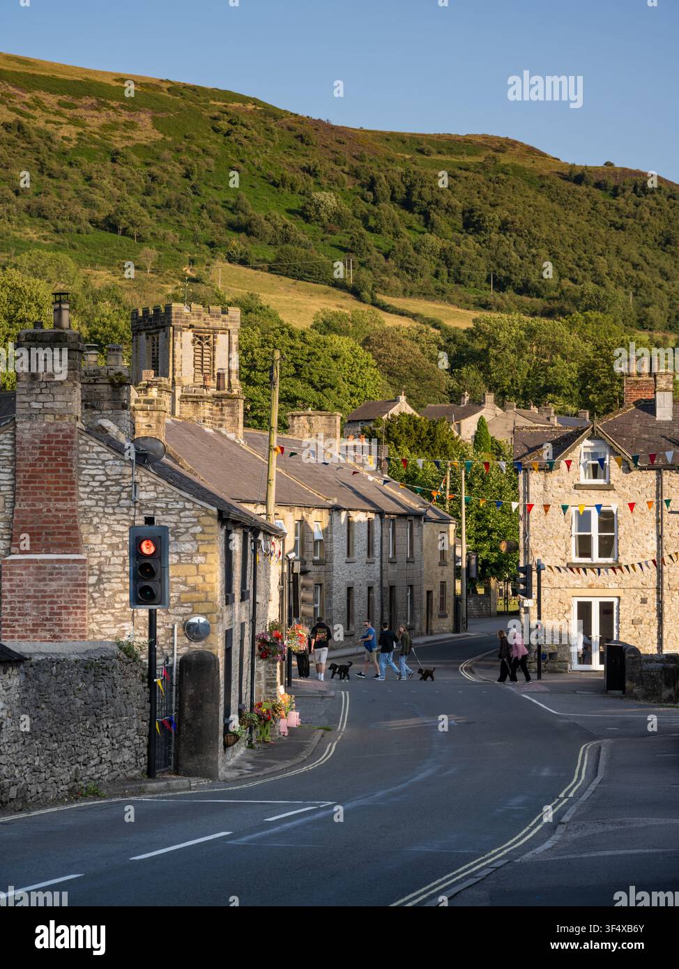 Menschen führen Hunde an einer festlich geschmückten Straße entlang, die mit historischen Steinhäusern und Fahnen geschmückt ist, im Dorfzentrum von Bradwell, Peak District, England Stockfoto