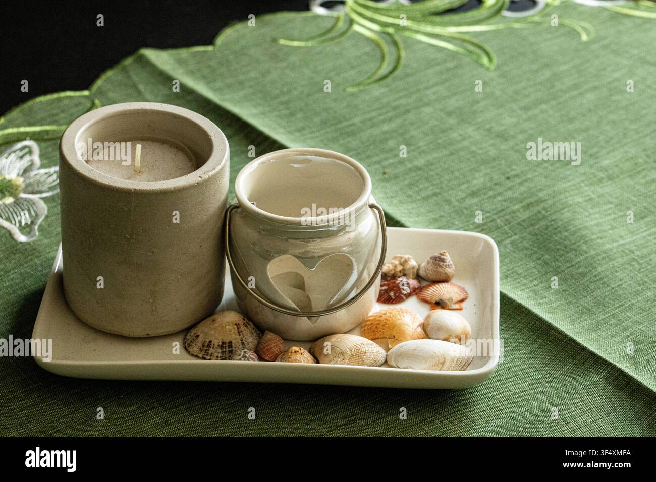 Dekorative Kerzen mit Muscheln und Kieseln, die auf einem Tablett über einer grünen Tischdecke arrangiert sind. Entspannende Spa- und Wellness-Stillleben. Stockfoto
