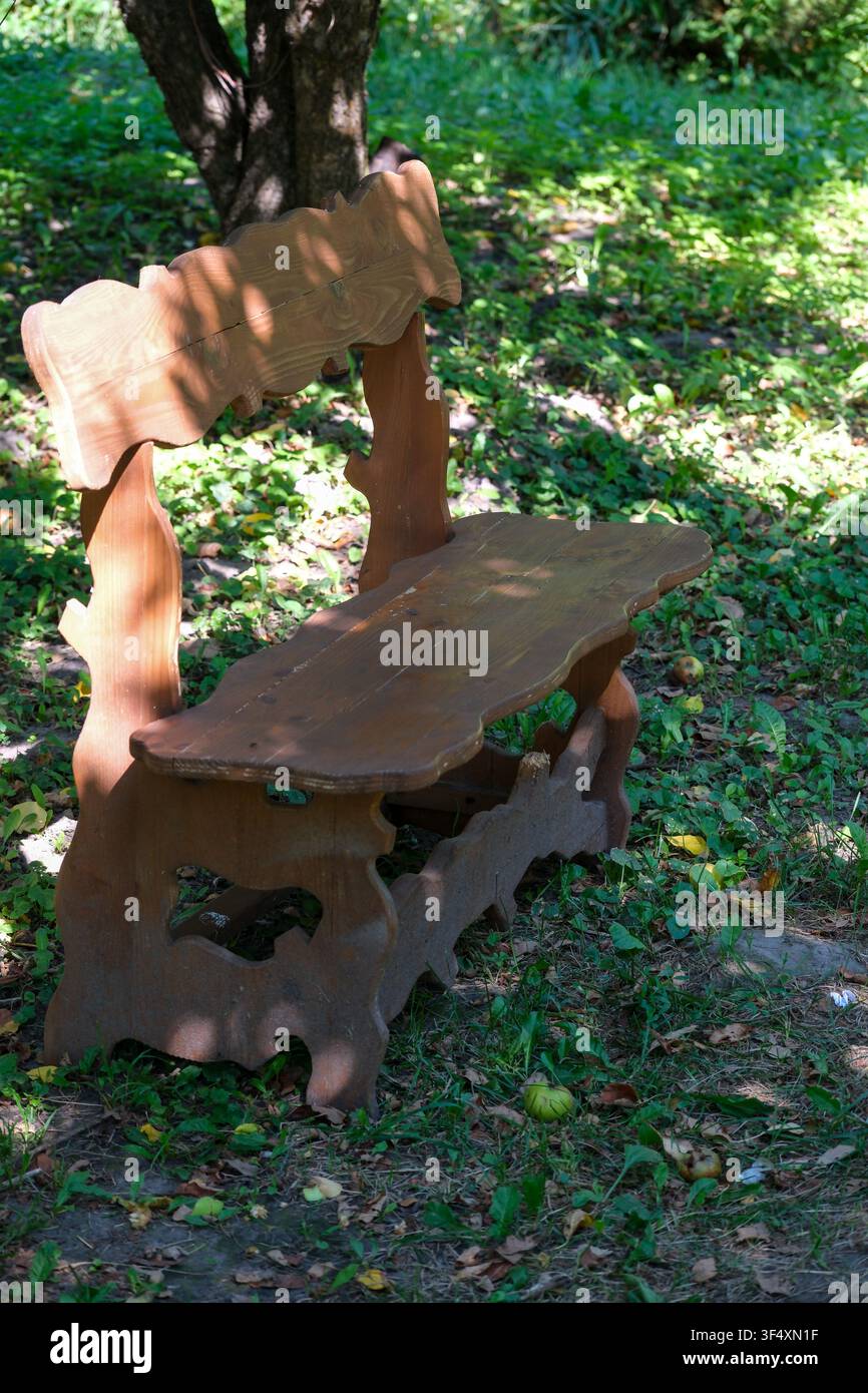 Rustische Holzgartenbank, die unter laubenden Bäumen in einem friedlichen Park an einem sonnigen Sommertag steht. Traditionelle Außensitzgelegenheit, umgeben von grünem Gras Stockfoto
