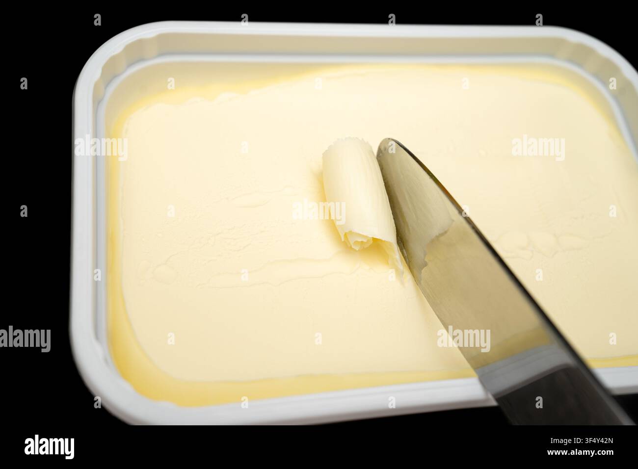 Ein Messer hebt vorsichtig ein kleines Stück Butter aus einem weißen Behälter. Die Küche ist einfach und funktional. Die Butter ist glatt und leicht zu verteilen. Stockfoto