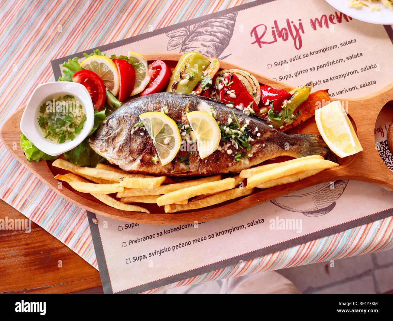 gegrillter Fisch mit Beilagen auf einem Holzteller, im Hintergrund eine Fischkarte "Riblji Menu" in südslawischer Sprache Stockfoto