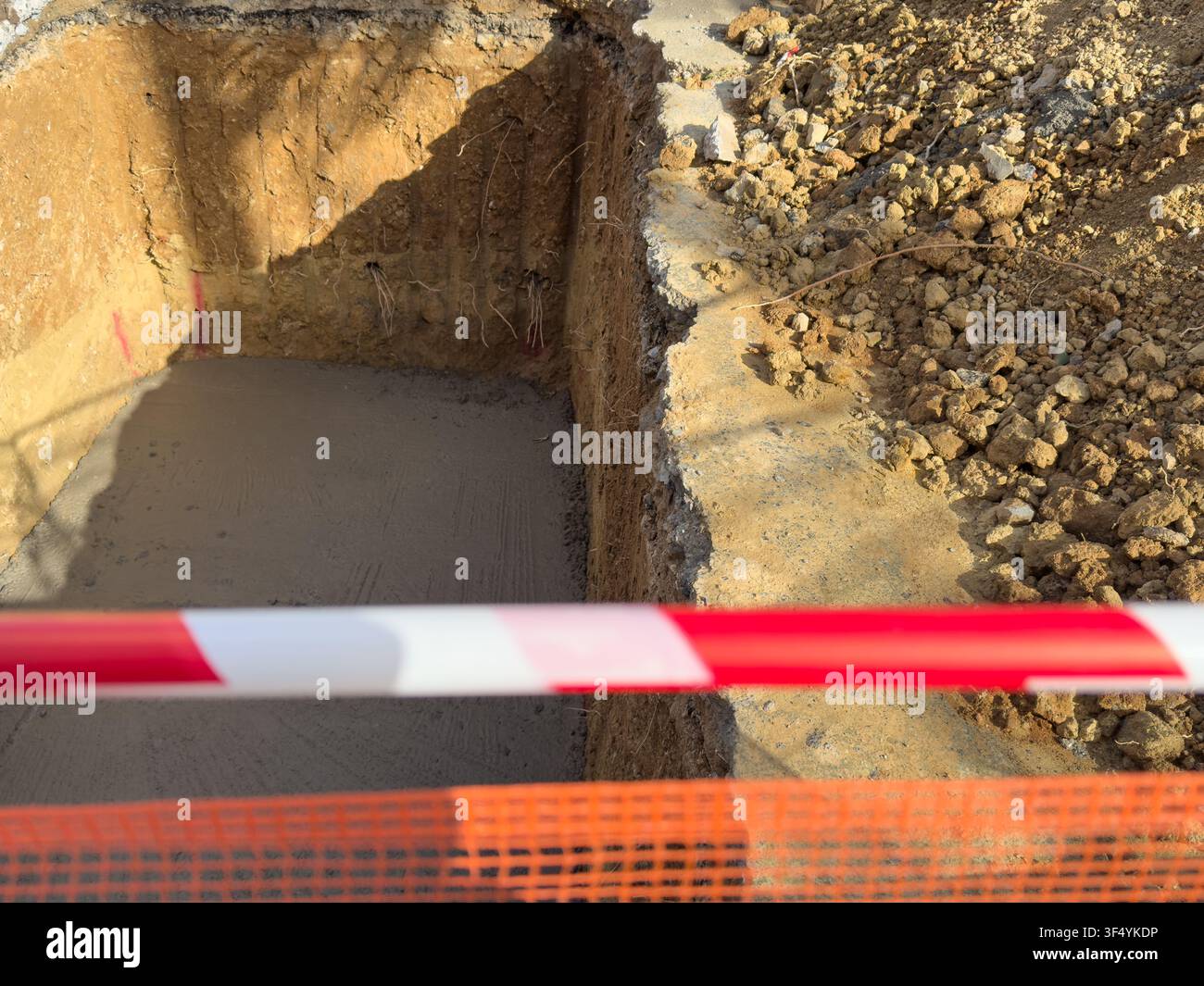 Tiefe Baugrube mit einem frisch gegossenen glatten Betonboden, begrenzt von rauer Erde und losem Kies, mit verschwommenem rot-weißem Sicherheitsband. Stockfoto