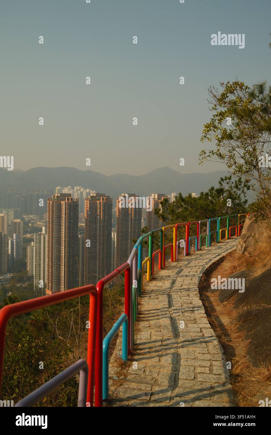 Vertikale Aufnahme der farbenfrohen Regenbogen-Geländer entlang des malerischen gepflasterten Bergpfades des Tuen Mun Trails mit Blick auf Hochhäuser in Hongkong. Stockfoto