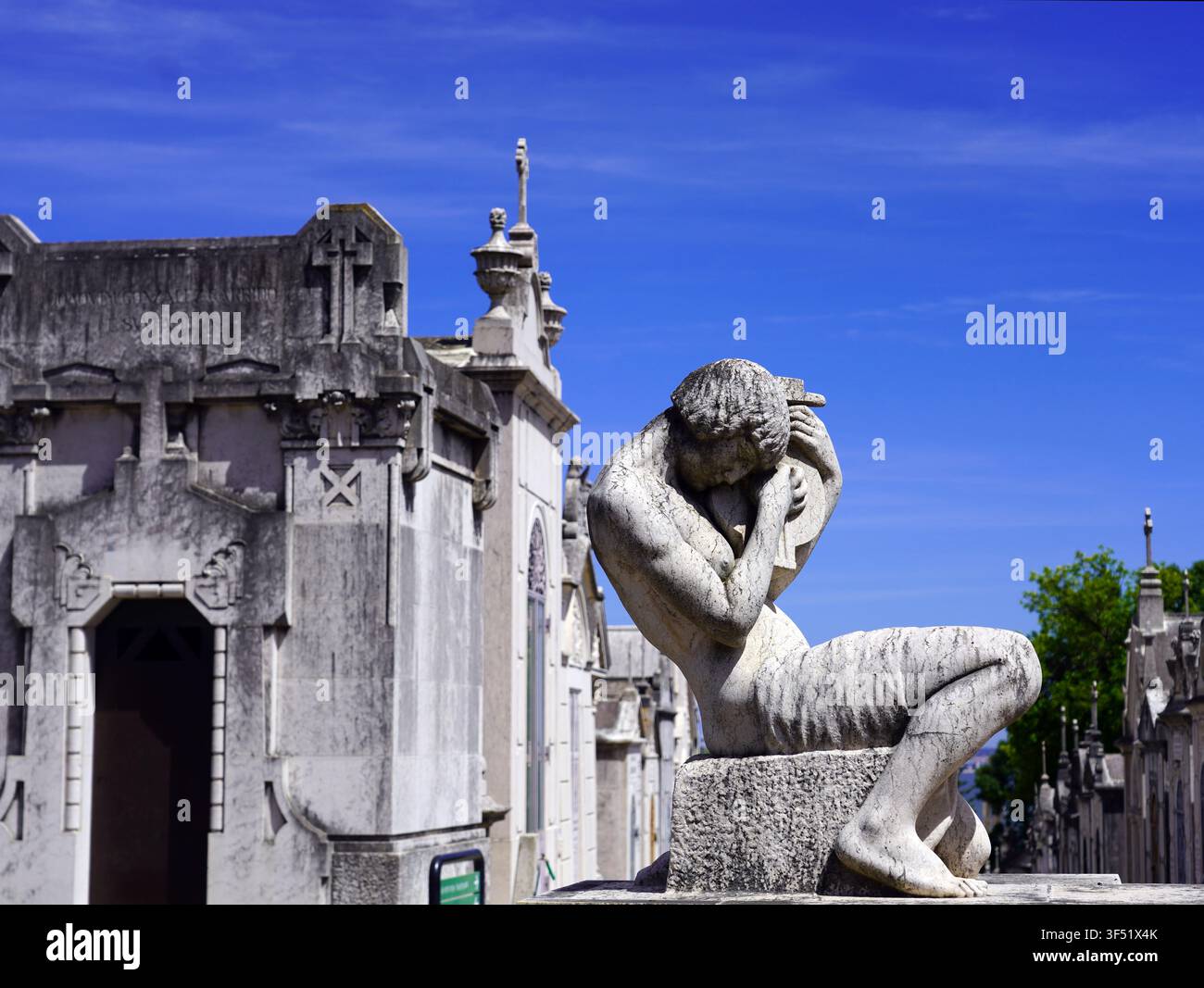Eine geschnitzte Steinstatue einer trauernden Gestalt steht neben historischen Mausoleen unter einem lebendigen blauen Himmel im Friedhof von Prazeres in Lissabon, Portugal Stockfoto