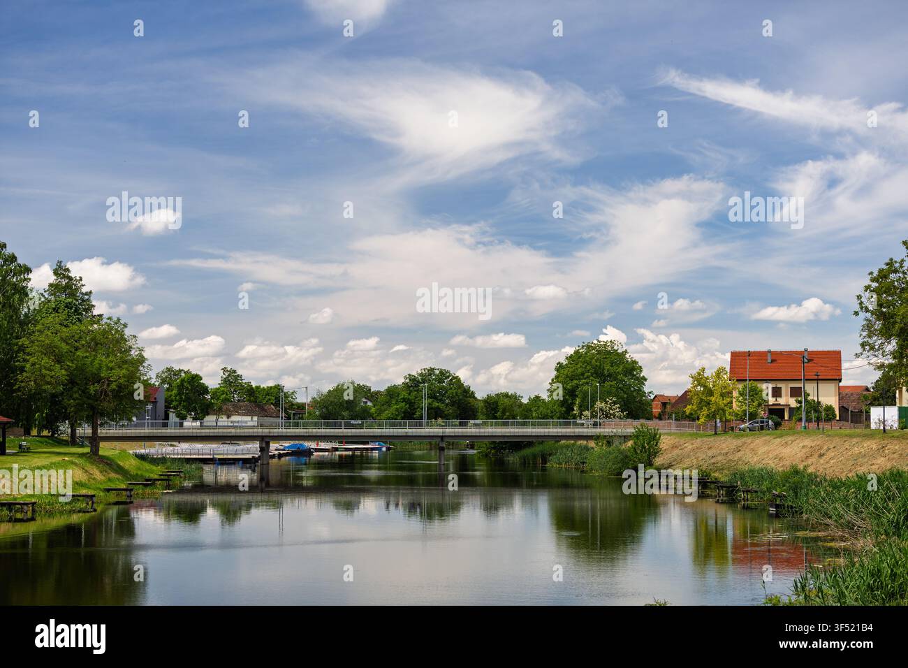 Friedliche Dorfszene mit einem kleinen Fluss mit Spiegelungen, einer niedrigen Betonbrücke, rot gedeckten Häusern und üppigen grünen Bäumen unter einem blauen Himmel mit Klo Stockfoto