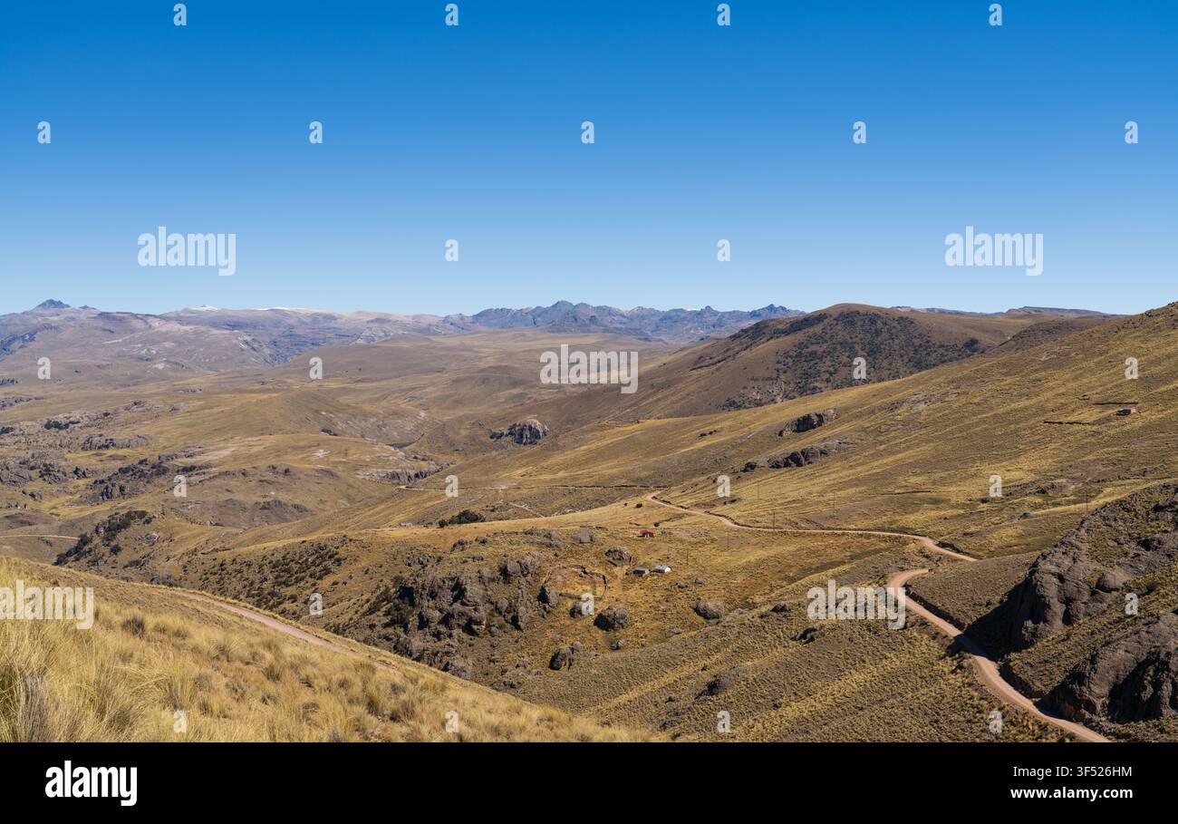 Panoramablick auf eine sich windende Schotterstraße, die sich durch die weite, trockene Bergtal-Landschaft des peruanischen Altiplano unter einem klaren blauen Himmel schlängelt Stockfoto