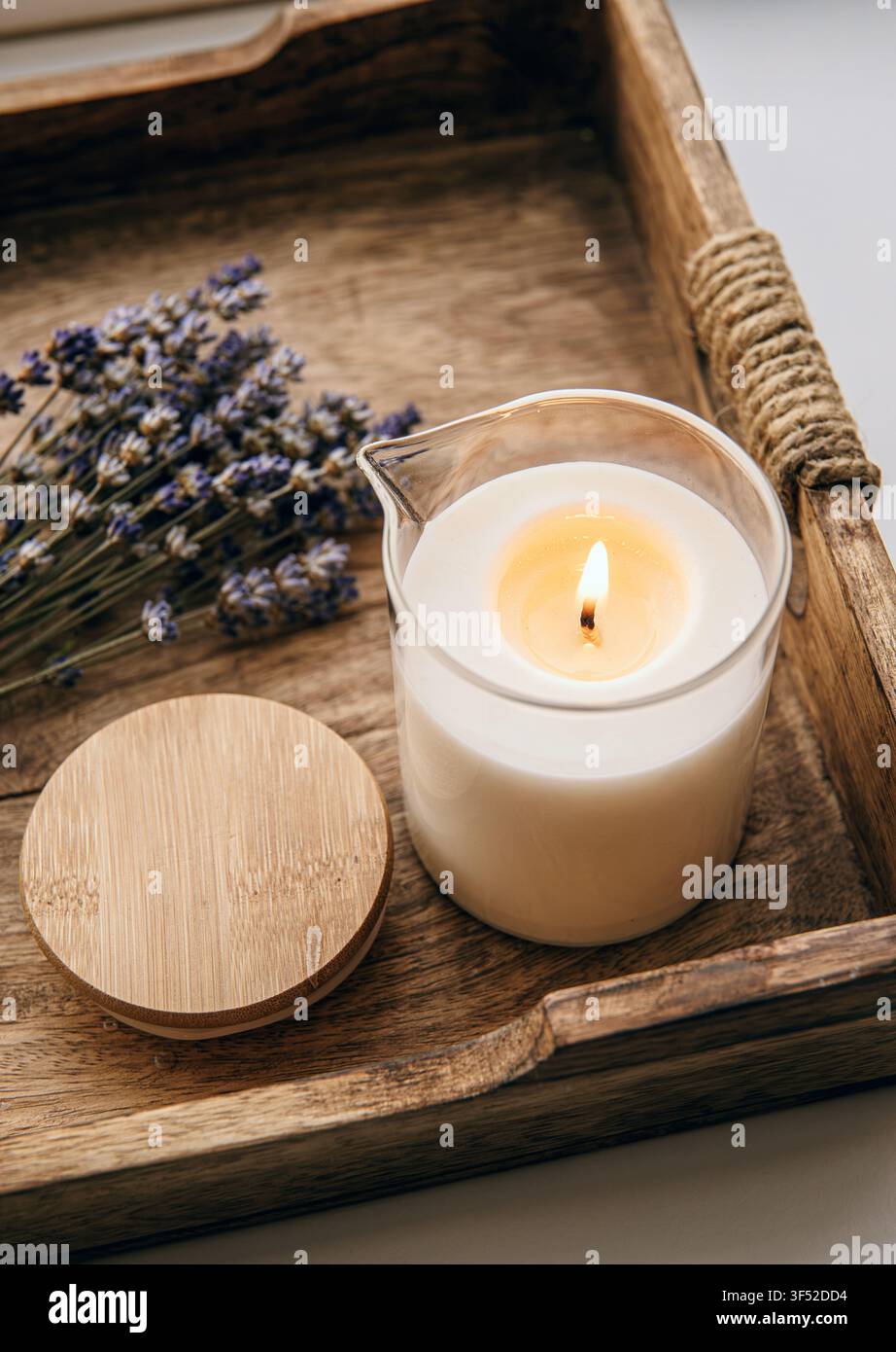 Eine selbstgemachte Massagekerze in einem Glasbehälter mit Ausguss, auf einem Holztablett platziert und mit einem Strauß getrockneter Lavendel dekoriert, drinnen zu Hause. Stockfoto