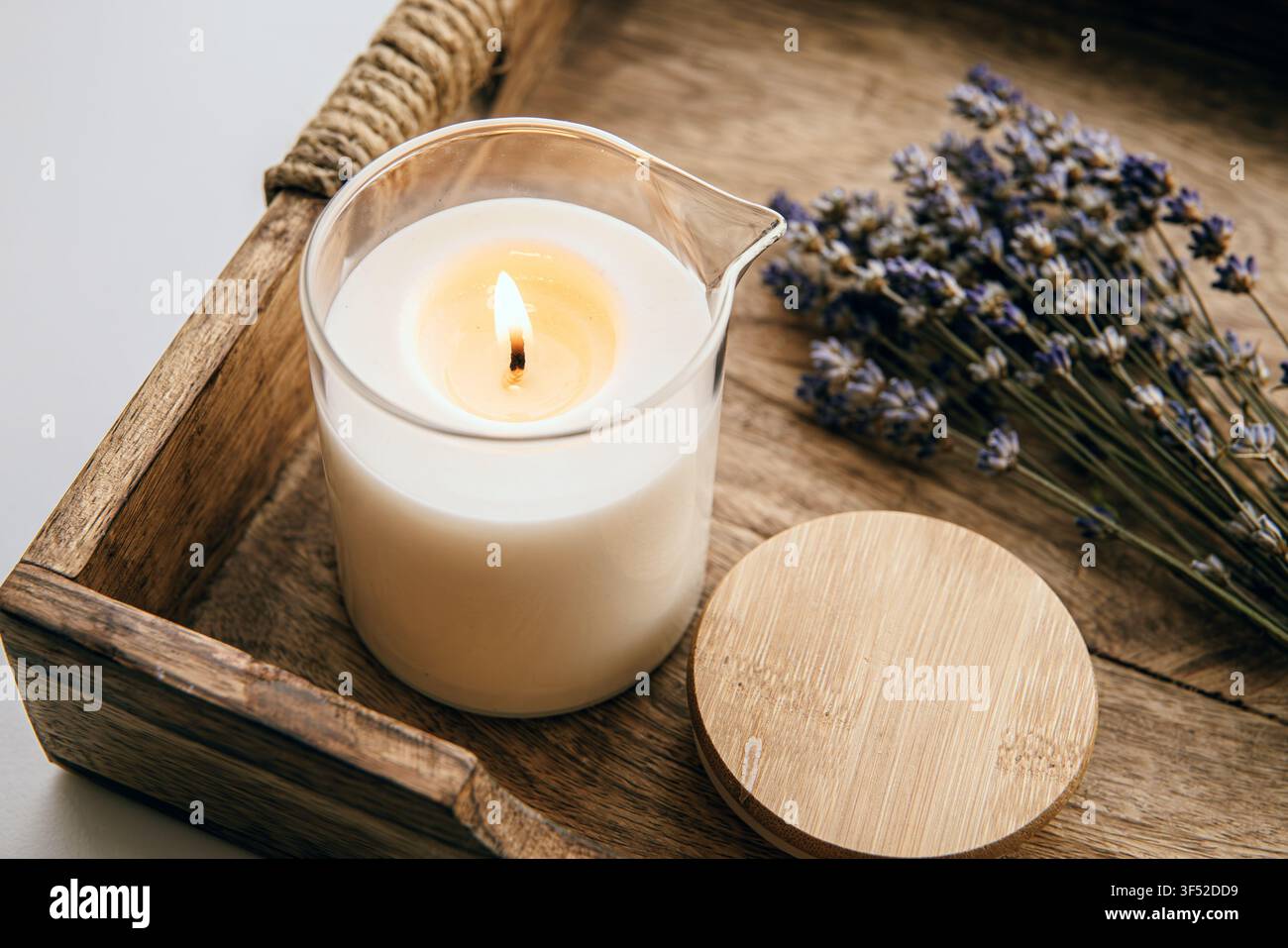 Eine selbstgemachte Massagekerze in einem Glasbehälter mit Ausguss, auf einem Holztablett platziert und mit einem Strauß getrockneter Lavendel dekoriert, drinnen zu Hause. Stockfoto