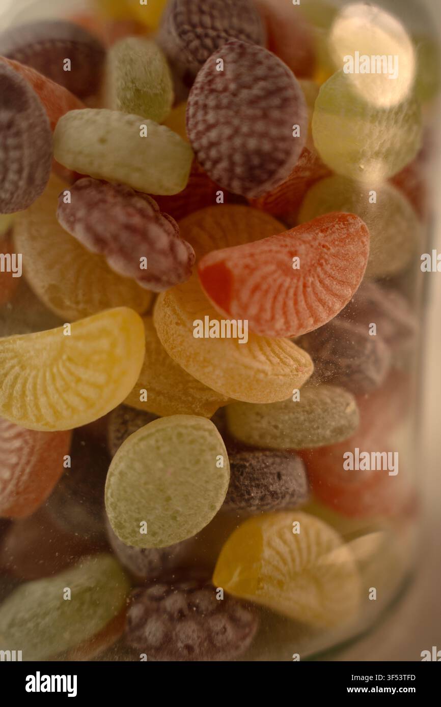 Makro-Nahaufnahme von verschiedenen gefrosteten, fruchtförmigen Hartbonbons, die in einem Glasgefäß gestapelt sind; warme Pastelltöne in Gelb, Orange, Grün und Lila, weiche flache Schärfentiefe Stockfoto