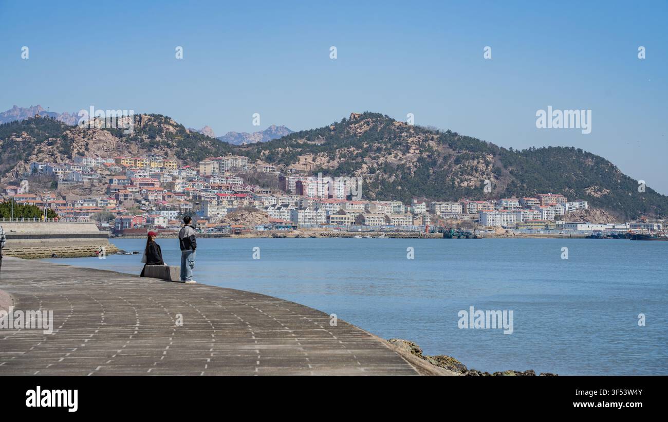Junges Paar entspannt sich auf einer Mole mit Blick auf das Küstendorf und die Hügel, Shazikou, Qingdao Stockfoto