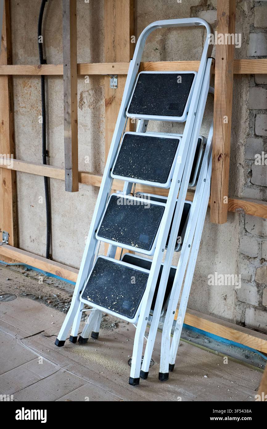 Weiße Metall-Klappleiter mit 4 Stufen steht nahe einer Holzkonstruktion auf staubigem Betonboden eines renovierten Hauses. Gebrauchte tragbare Ausrüstung mit rutschfester S Stockfoto