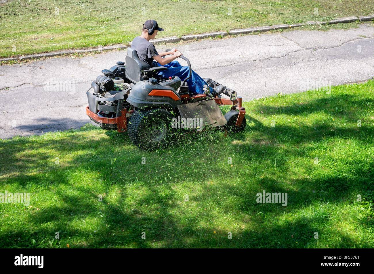 Gartenpfleger fährt einen Zero-Turn-Rasenmäher im öffentlichen Park. Kommunaler Rasenmähertraktor schneidet grünes Gras entlang der Straße mit fliegenden Grashalmen Stockfoto