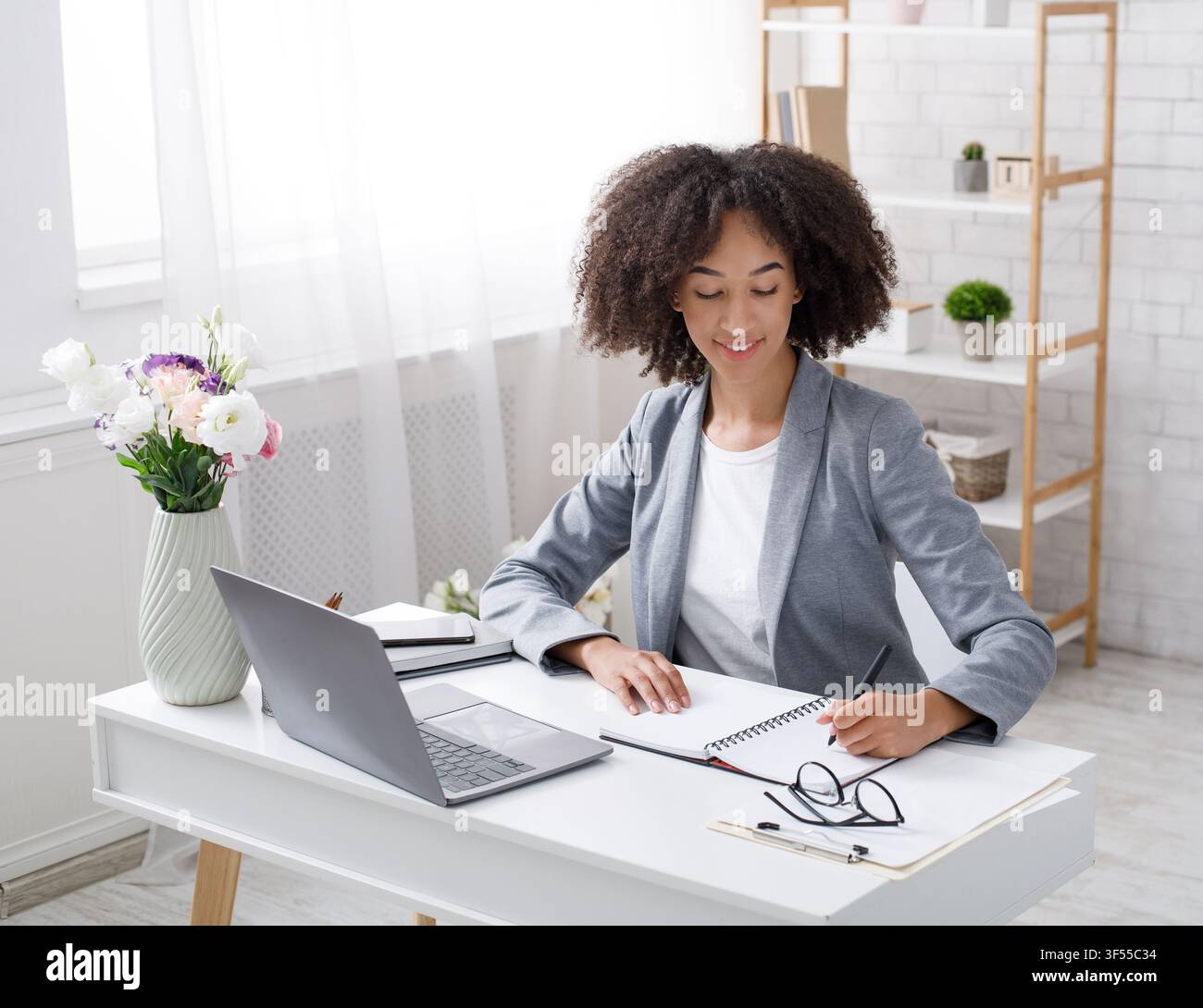 Nehmen Sie während des Ausbruchs eine Bestellung von Kunden im Online-Büro zu Hause an. Fröhliche afroamerikanische Managerin arbeitet am Tisch mit Laptop und schreibt Stockfoto