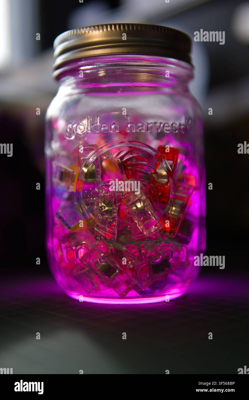Ein Mason-Glas der Marke Golden Harvest, das in rosa Licht leuchtet, gefüllt mit klaren Plastikgegenständen, vor dunklem Hintergrund. Stockfoto