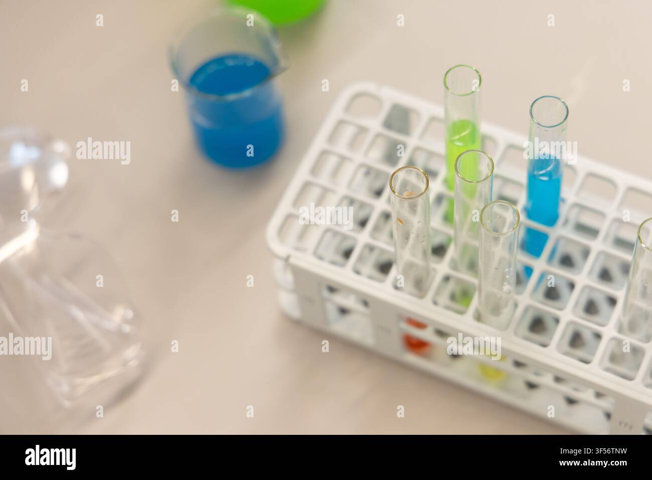 Reagenzglasgestell mit Glasröhrchen mit blauen und grünen Flüssigkeiten auf beiger Arbeitsplatte, unscharfer Messbecher Stockfoto