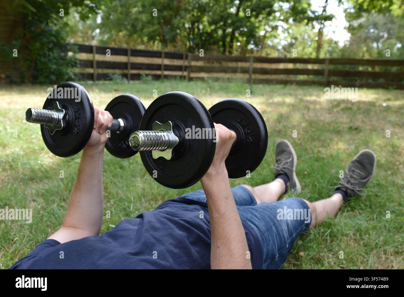 Zwei Hände einer Person, jede von ihnen hält ein Hantelbär. Die Hanteln werden in die Luft gehoben. Grünes Gras, Büsche, ein Holzzaun im Hintergrund. Stockfoto