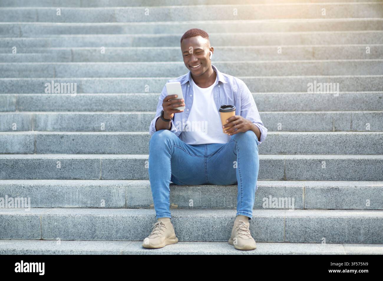 Urbaner Lebensstil. Attraktiver schwarzer Mann mit Kaffee zum Mitnehmen mit Handy auf Steintreppen draußen Stockfoto