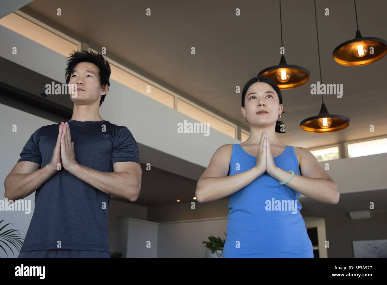 Asiatisches Paar steht in einem modernen Studio mit Pendelleuchten und drückt die Handflächen an der Brust in Sportkleidung Stockfoto