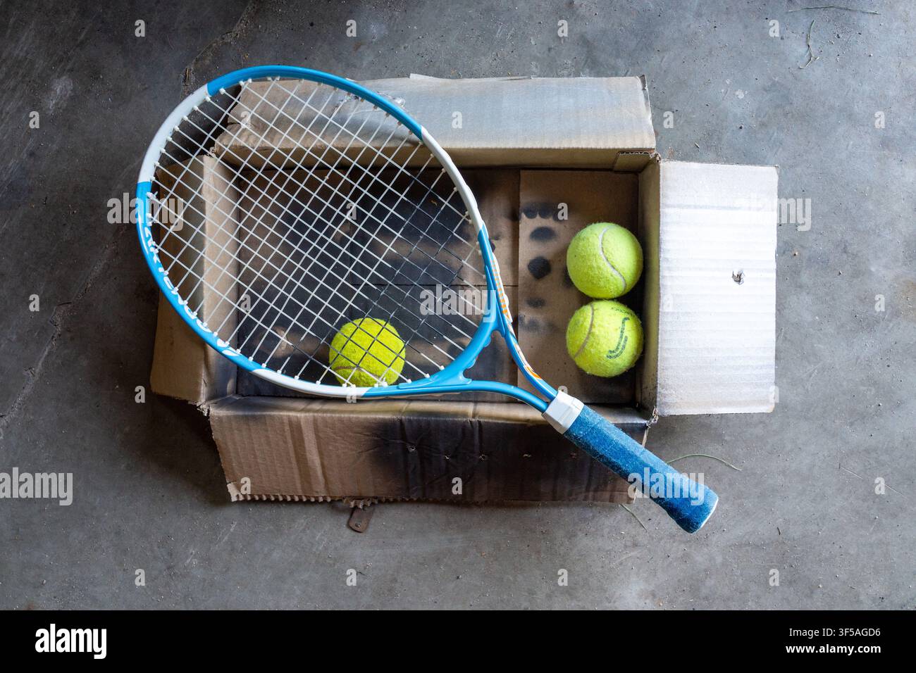 Ansicht von oben eines blauen Tennisschlägers und gelber Bälle in einer offenen Pappschachtel auf rauem grauem Beton, zeigt Sportverpackung, Übung, Lieferung und Lagerkonzept für den Einzelhandel Stockfoto