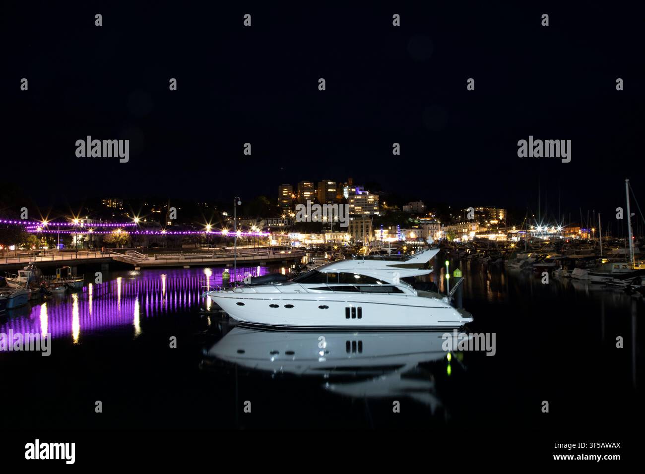 Ein luxuriöses Motorboot ruht auf ruhigem Wasser und wirft nachts ein scharfes Spiegelbild im Hafen von Torquay neben der beleuchteten Millennium Bridge. Stockfoto