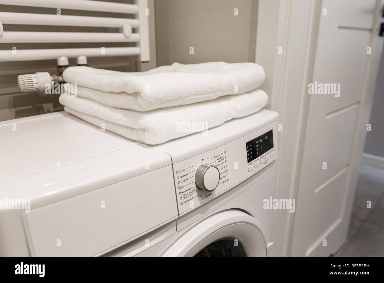 Stapel frischer sauberer weißer gefalteter Handtücher oben auf einer modernen weißen Waschmaschine in einem Badezimmerambiente. Organisation von Haushaltswaschräumen, Textilhygiene, minimalistische Innenausstattung Stockfoto