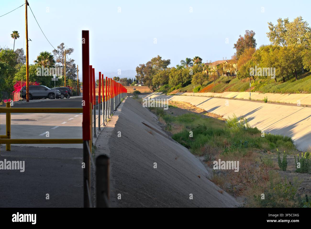 Ein trockener Beton-Überwasserkanal verläuft neben einem Parkplatz und einem landschaftlich gestalteten Hügel, gesäumt von Barrieren, Unkraut, Bäumen und nahe gelegenen Häusern. Stockfoto