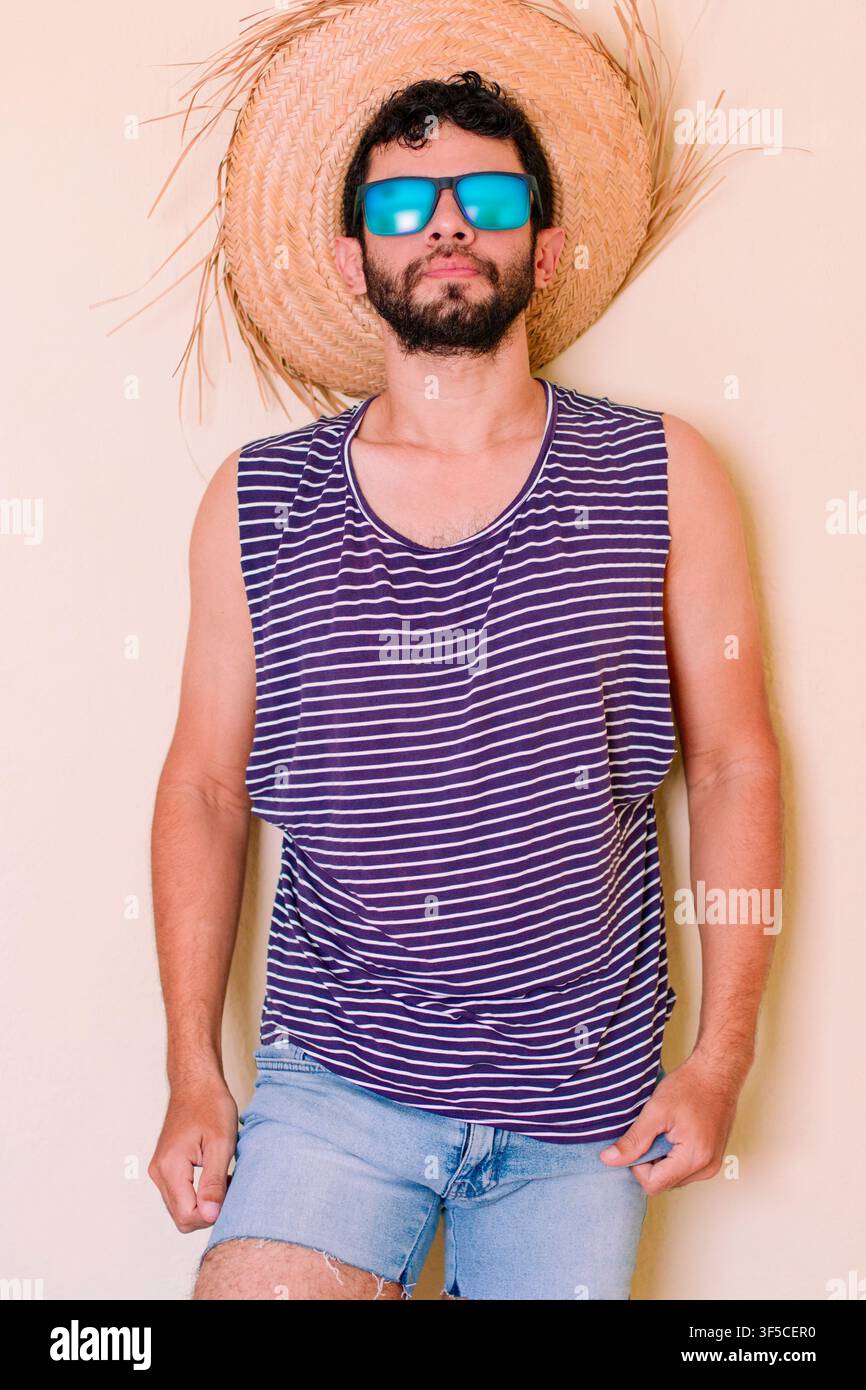 Tourist mit Brille und Hut. Junger männlicher Tourist trägt ein gestreiftes Tanktop im Matrosenstil und kurze Jeansshorts. Er spürt die Sommerhitze Stockfoto