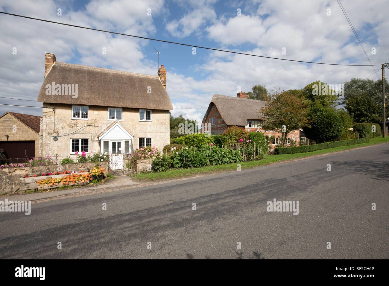 Traditionelle strohgedeckte Steinhäuser mit gepflegten Vorgärten stehen an einer ländlichen Dorfstraße unter einem blauen Himmel in der englischen Landschaft. Stockfoto