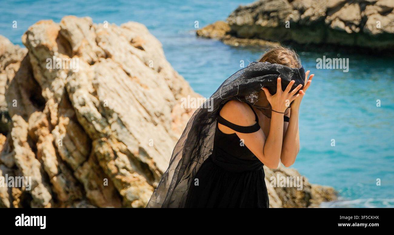 Junge Frau in schwarzem Kleid mit schwarzem Schal, die auf den Felsen steht Stockfoto