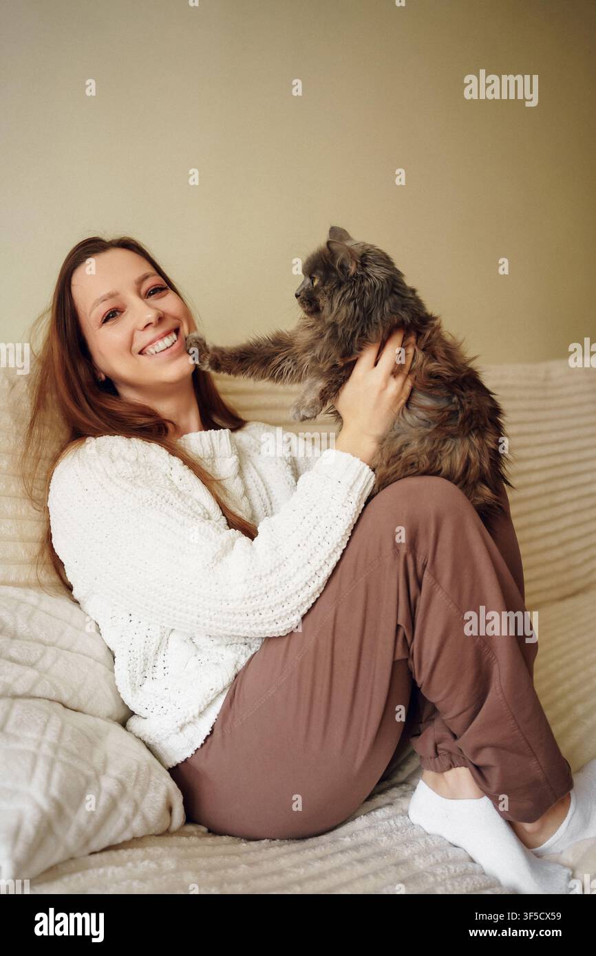 Ein warmes und intimes Porträt einer fröhlichen jungen Frau mit langen Haaren, die bequem auf einer weichen Couch zu Hause sitzt. Sie trägt einen strukturierten cremeweißen Pullover und braune Lounge-Hosen und ist glücklich Stockfoto