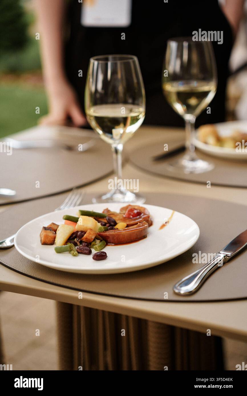 Gourmet-Mahlzeit auf einem weißen Teller mit Gläsern weißer Wein auf einem Tischgedeck, perfekt für Restaurant-Marketing, Food-Blogs oder Catering-Events, Esserlebnis und feines Catering Stockfoto