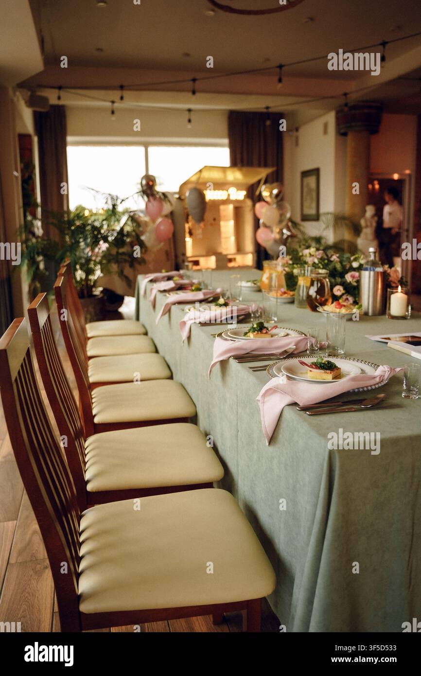 Langer Esstisch, der für eine formelle Veranstaltung mit salbeigrünen Tischdecken, rosafarbenen Servietten und festlicher Dekoration in einem gemütlichen Restaurantinterieur arrangiert ist, festliche Dekoration Stockfoto