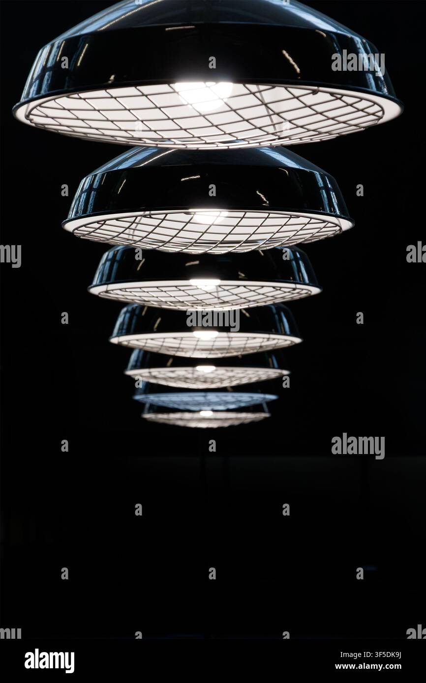 Reihe von beleuchteten Pendelleuchten im Industrie-Stil mit Metallgitterabdeckungen, die in Perspektive vor einem dunklen Hintergrund hängen Stockfoto