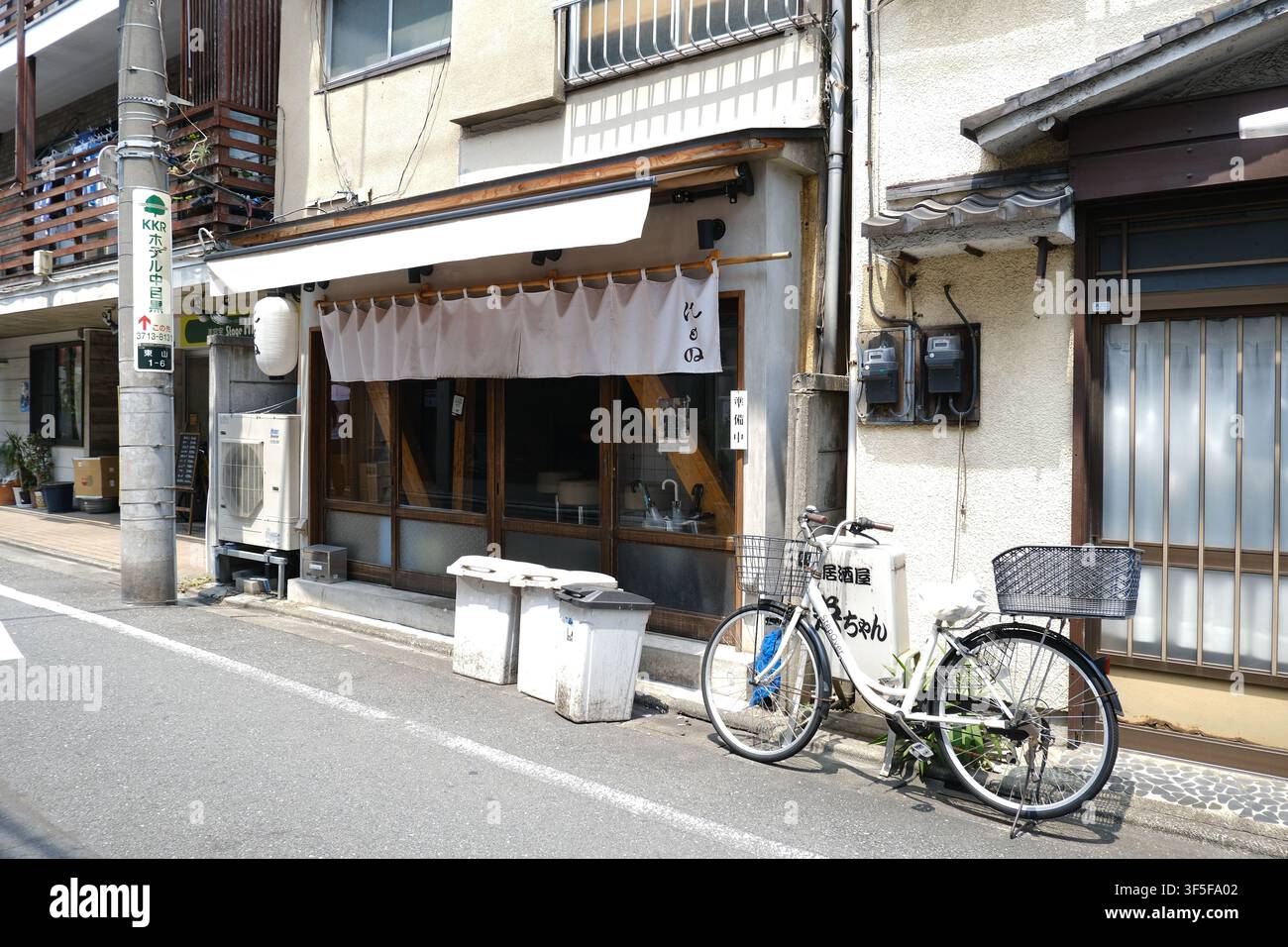 Ein weißes Fahrrad mit Vorder- und Hinterkorb, das vor einem japanischen Restaurant mit einem weißen Noren-Vorhang und Mülltonnen entlang einer Straße in Tokio geparkt ist Stockfoto