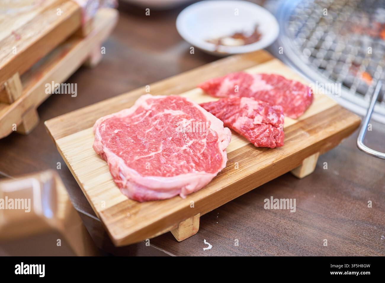 Verschiedene rohe koreanische Fleischstücke auf einem Holzbrett neben einem Holzkohlegrill, zubereitet für ein traditionelles koreanisches Barbecue-Essen. Stockfoto