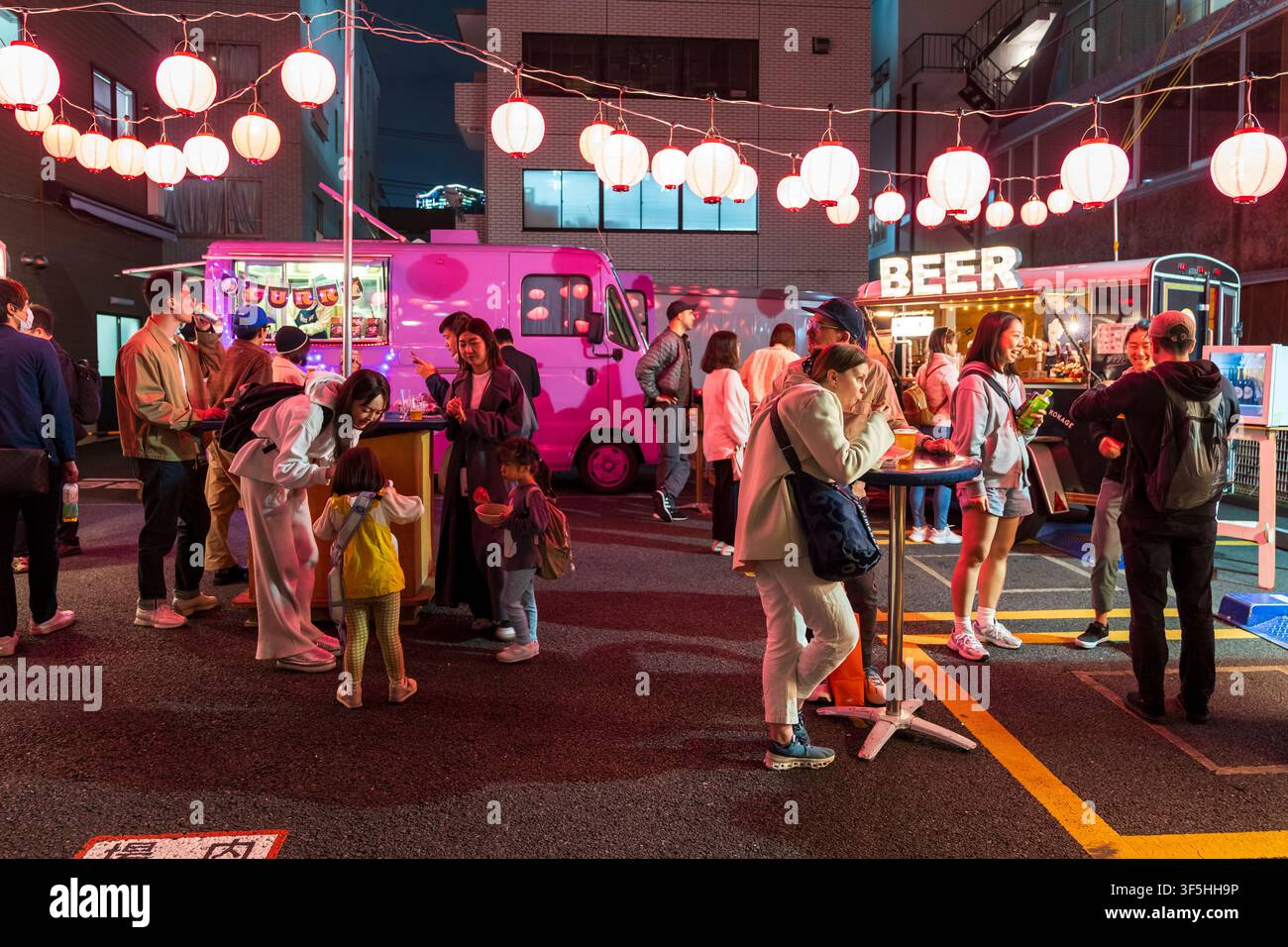 Tokio-Nachtfest mit Foodtrucks und Papierlaternen. Gruppen von Menschen und eine Familie genießen Essen und Getränke an Tischen, die auf einem Parkplatz aufgestellt sind. Stockfoto