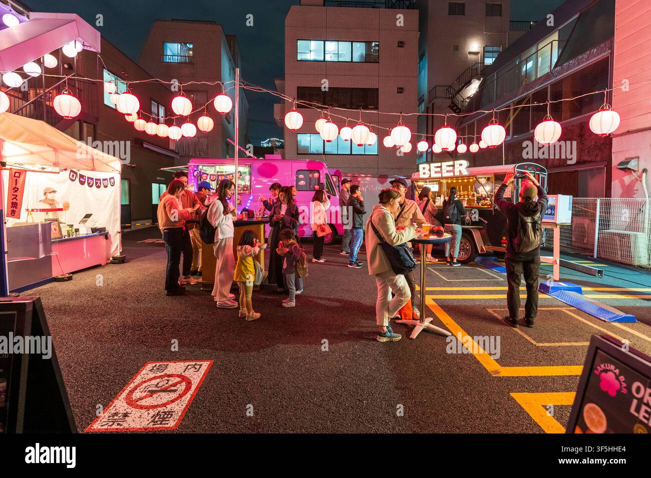 Tokio-Nachtfest mit Foodtrucks und Papierlaternen. Gruppen von Menschen und eine Familie genießen Essen und Getränke an Tischen, die auf einem Parkplatz aufgestellt sind. Stockfoto