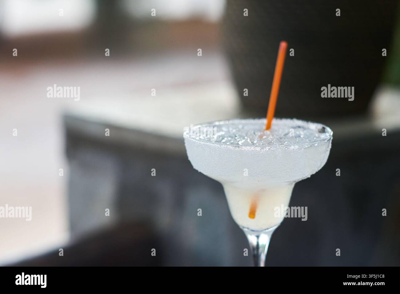 Nahaufnahme eines erfrischenden gefrorenen Margaritas-Cocktails, serviert in einem traditionellen Glas mit salziger Bordüre und einem Orangenstrohhalm, der auf einem Außentisch steht. Stockfoto