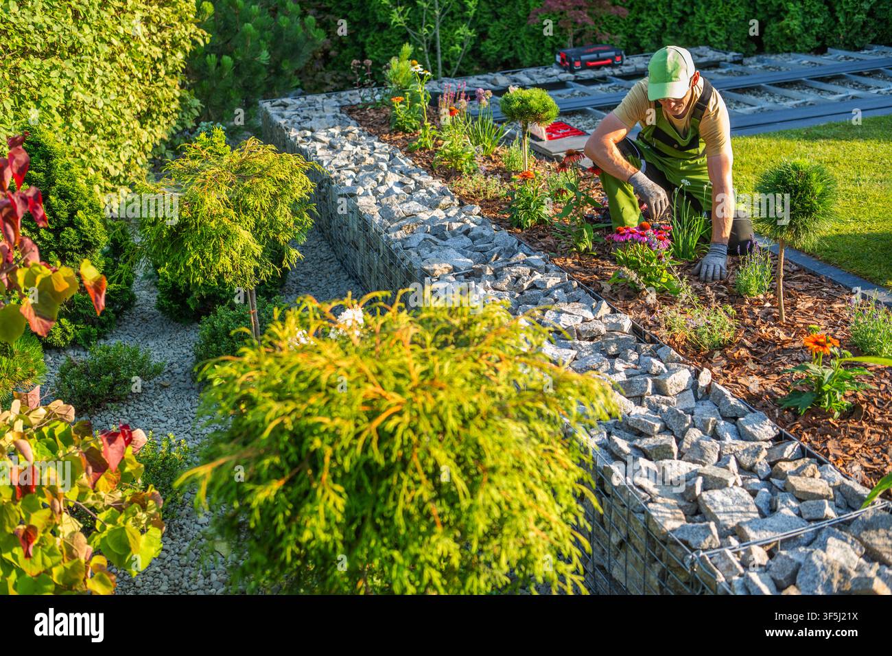 Ein Gärtner pflanzt Blumen in einem ordentlich angeordneten Garten an einem Wohngrundstück. Der Garten bietet Steine, Pflanzen und farbenfrohe Blumen. Der ga Stockfoto