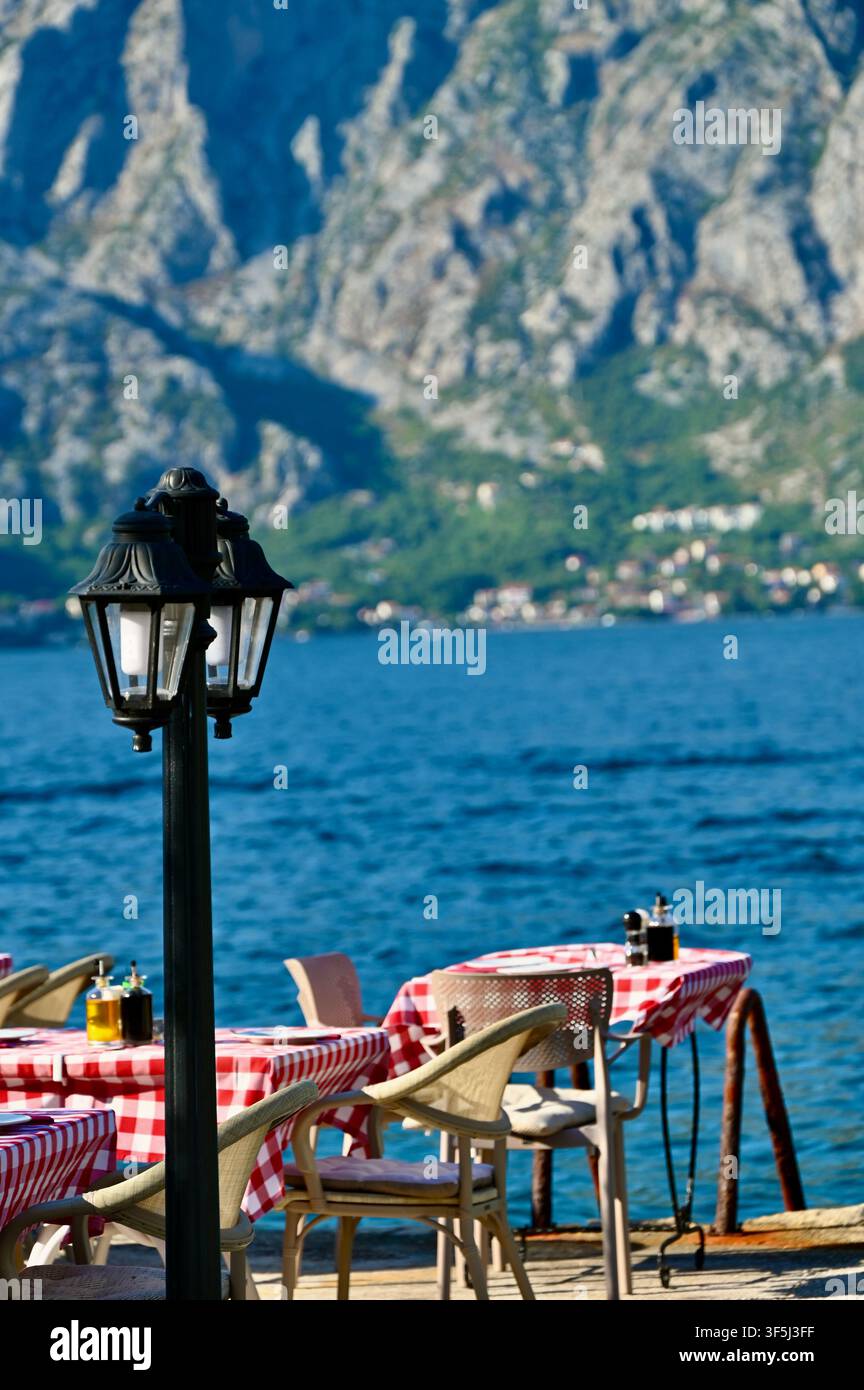 Eine Außenterrasse eines Cafés am blauen Meer mit einem schwarzen Vintage-Lampepfosten im Vordergrund, Tischen mit karierten Tischdecken und einer majestätischen Bergkulisse. Stockfoto
