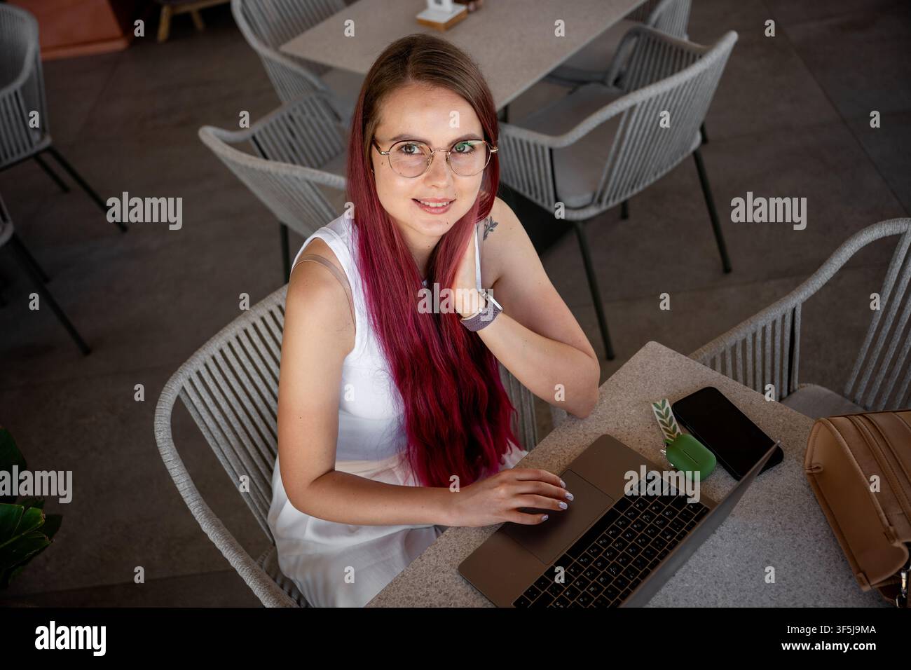 Ein moderner Freiberufler mit roten Haaren und Brille arbeitet an einem Laptop in einem modischen Café. Er genießt eine stilvolle, entspannte Umgebung und balanciert Produk Stockfoto